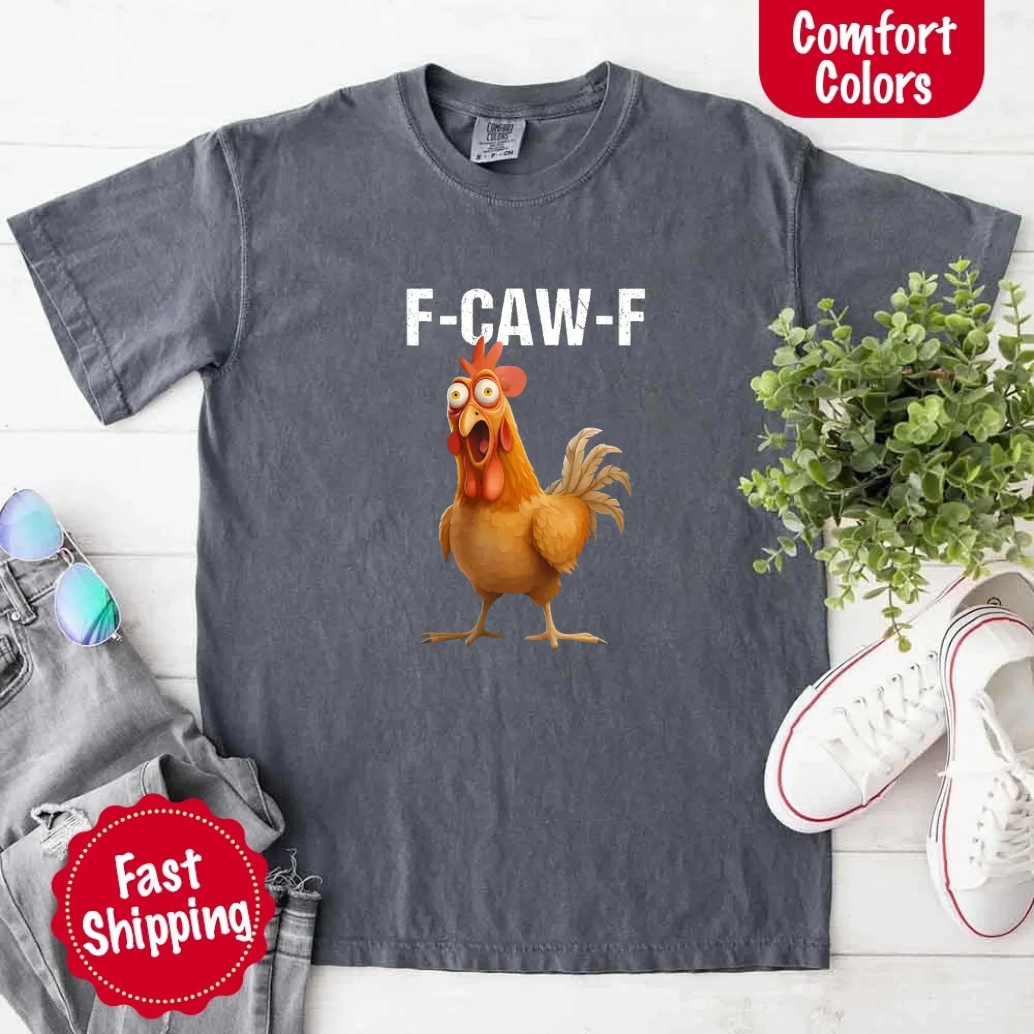 Gray t-shirt with a cartoon chicken and 'F-CAW-F' text on a white surface with accessories.