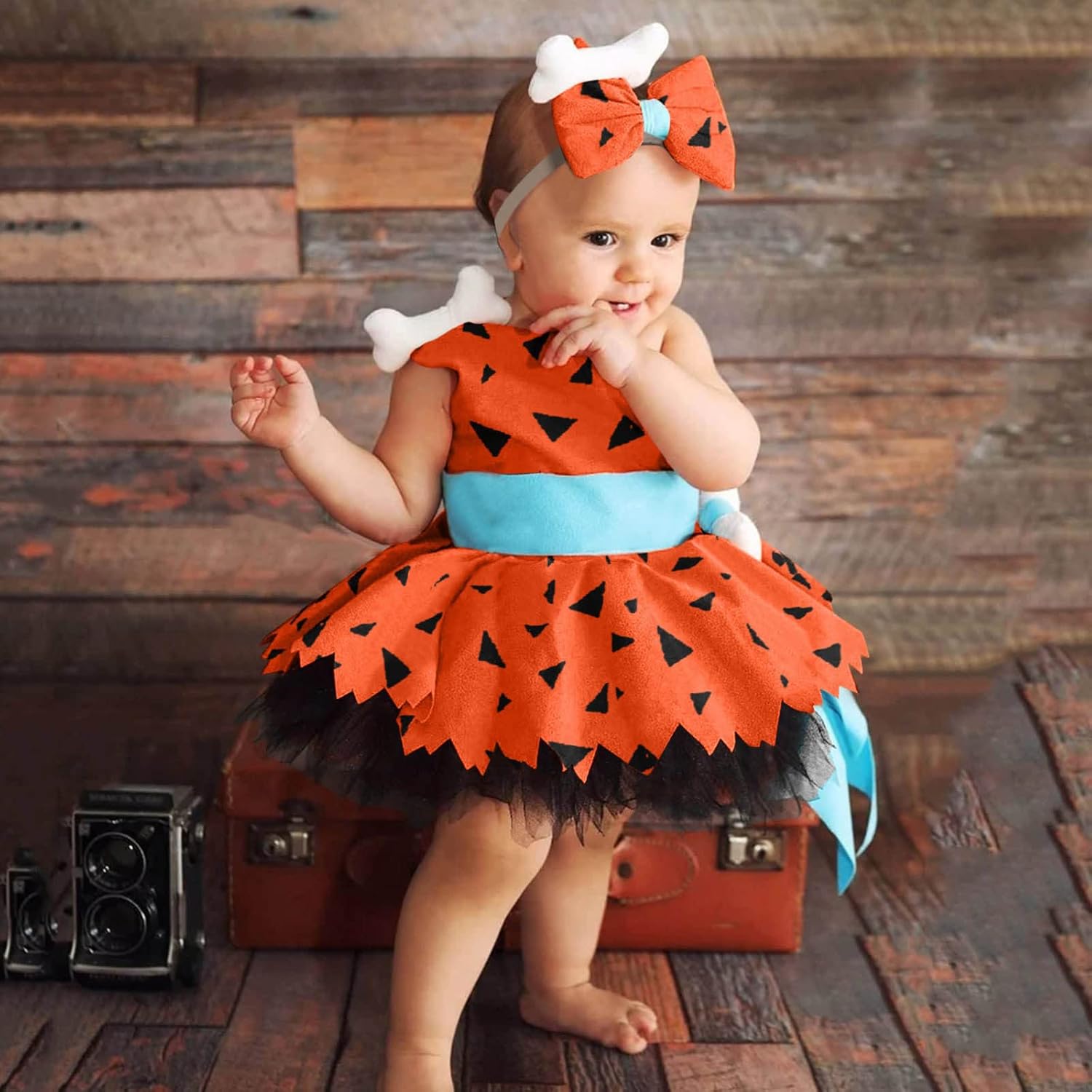 Baby in a Halloween-themed outfit with an orange dress and headband, sitting on a wooden floor.