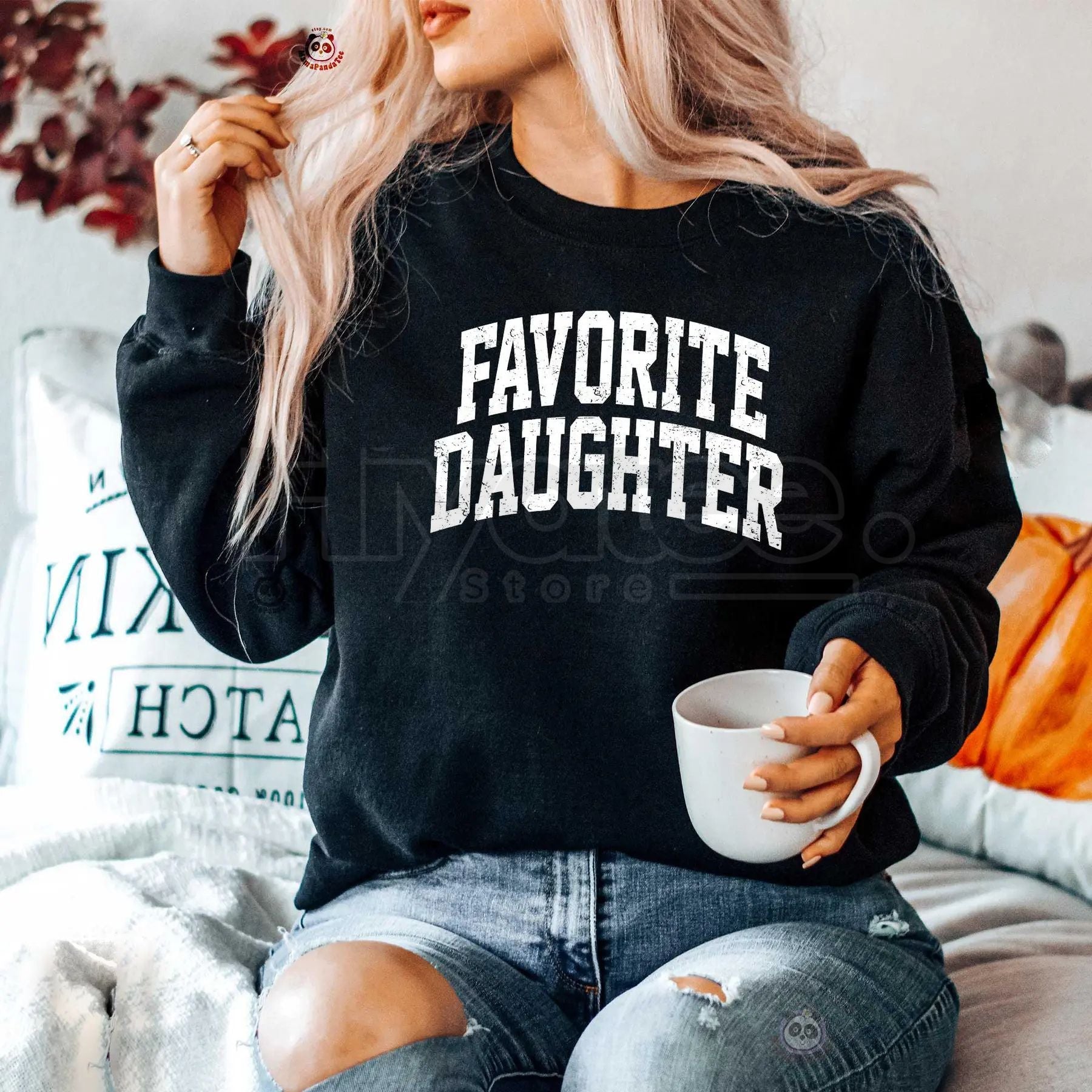 Person wearing a black sweatshirt with 'FAVORITE DAUGHTER' text, holding a mug.