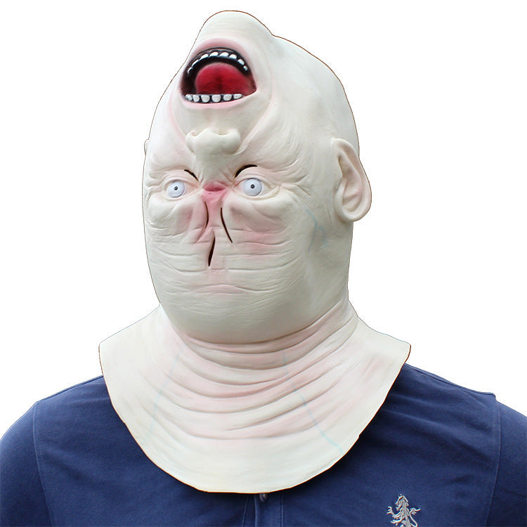 A pale, grotesque latex mask with an open mouth showing red interior and white teeth, blue eyes, and wrinkled skin, worn over a person's head and neck with a navy shirt visible below, on a plain white background.