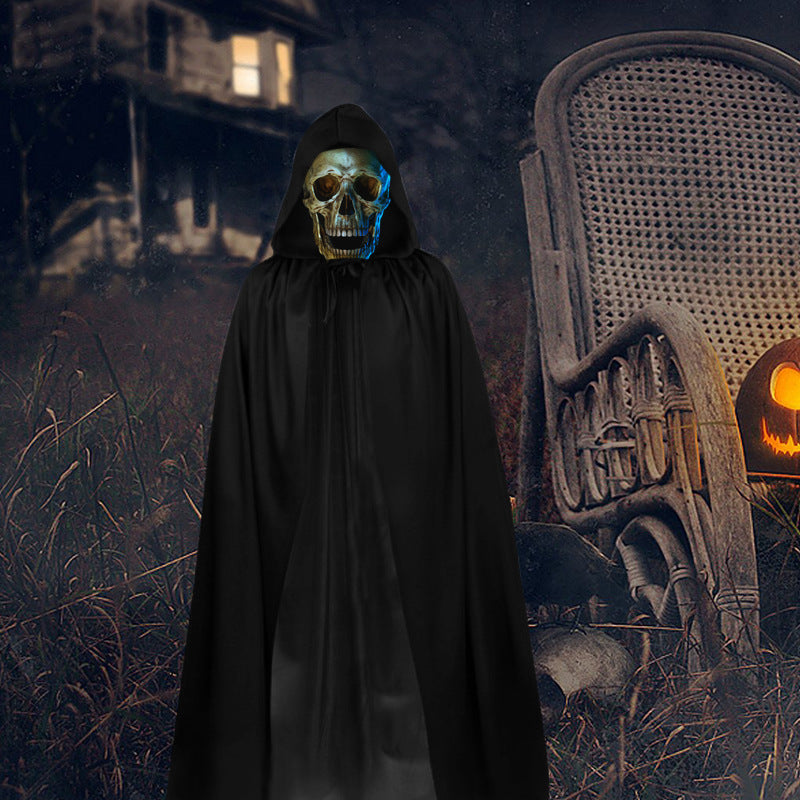 A person wearing a black hooded robe and a metallic silver skull mask stands in a dark outdoor Halloween scene with a chair and jack-o'-lantern in the background.