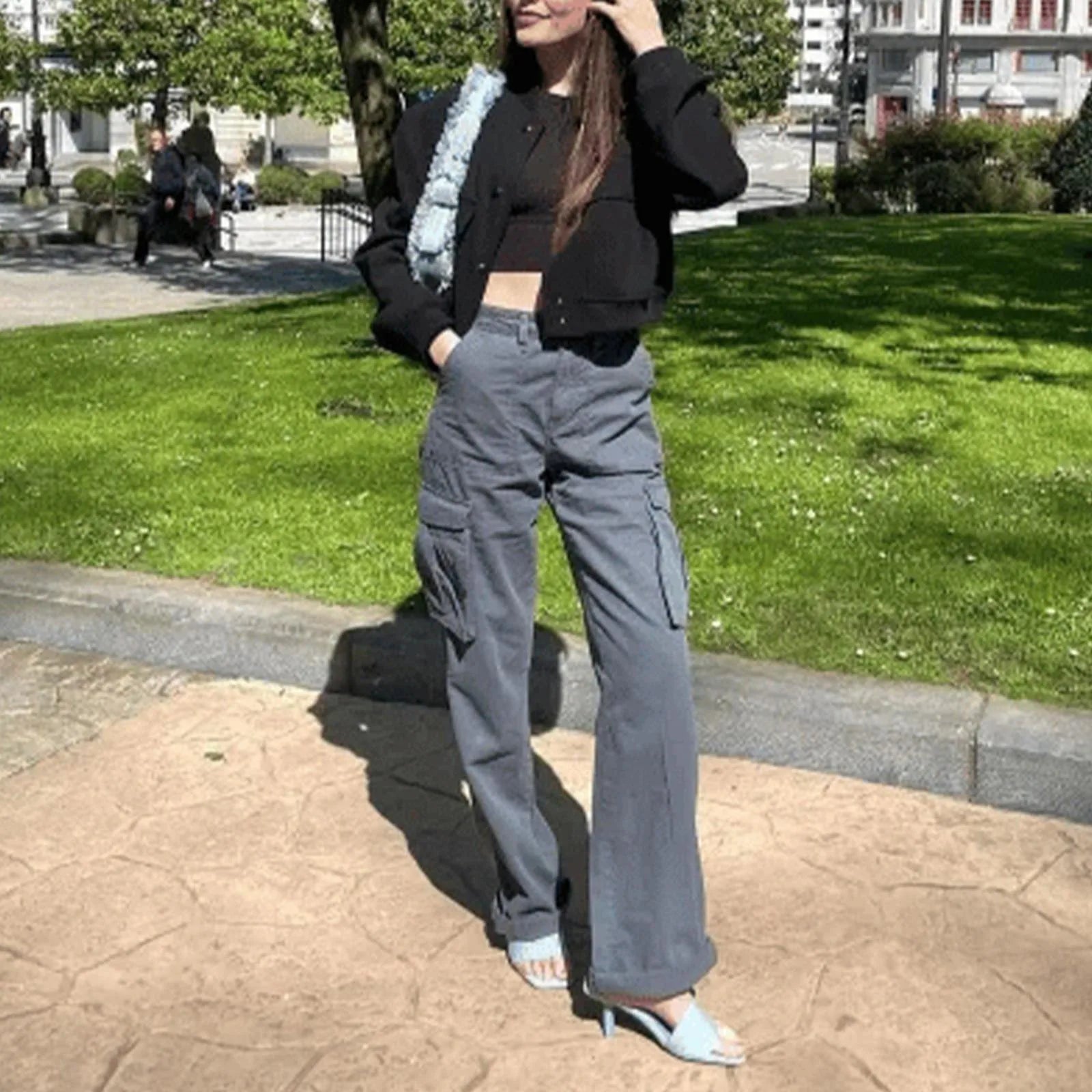 Person wearing a black jacket and gray cargo pants standing on a sidewalk with green grass and trees in the background.