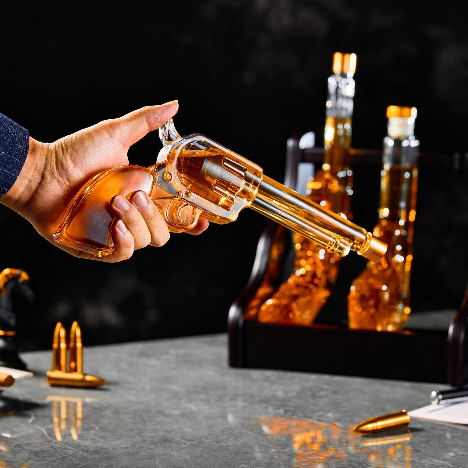 Hand holding a glass revolver with alcohol, surrounded by similar bottles and bullets on a dark surface.