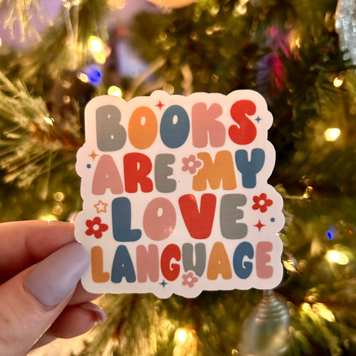 Colorful sticker with 'Books are my love language' text held in front of a decorated Christmas tree.