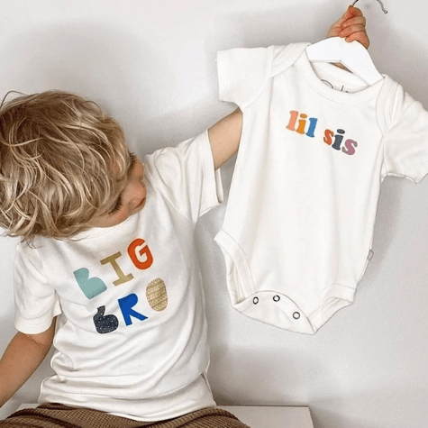 Child wearing a 'BIG BROTHER' shirt holding a 'lil sis' onesie on a hanger.