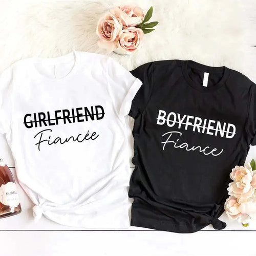 White and black t-shirts with 'Girlfriend Fiançe' and 'Boyfriend Fiançe' text on a white surface with flowers.