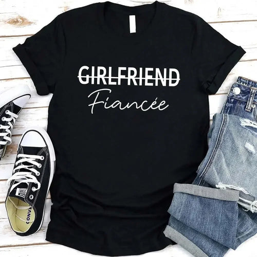 Black t-shirt with 'GIRLFRIEND Fiançée' text, paired with jeans and sneakers on a wooden surface.