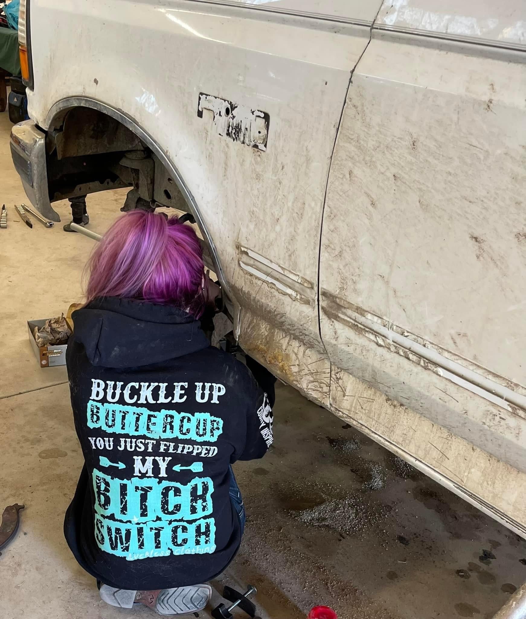 Person with purple hair working on a car in a garage, wearing a jacket with offensive text.