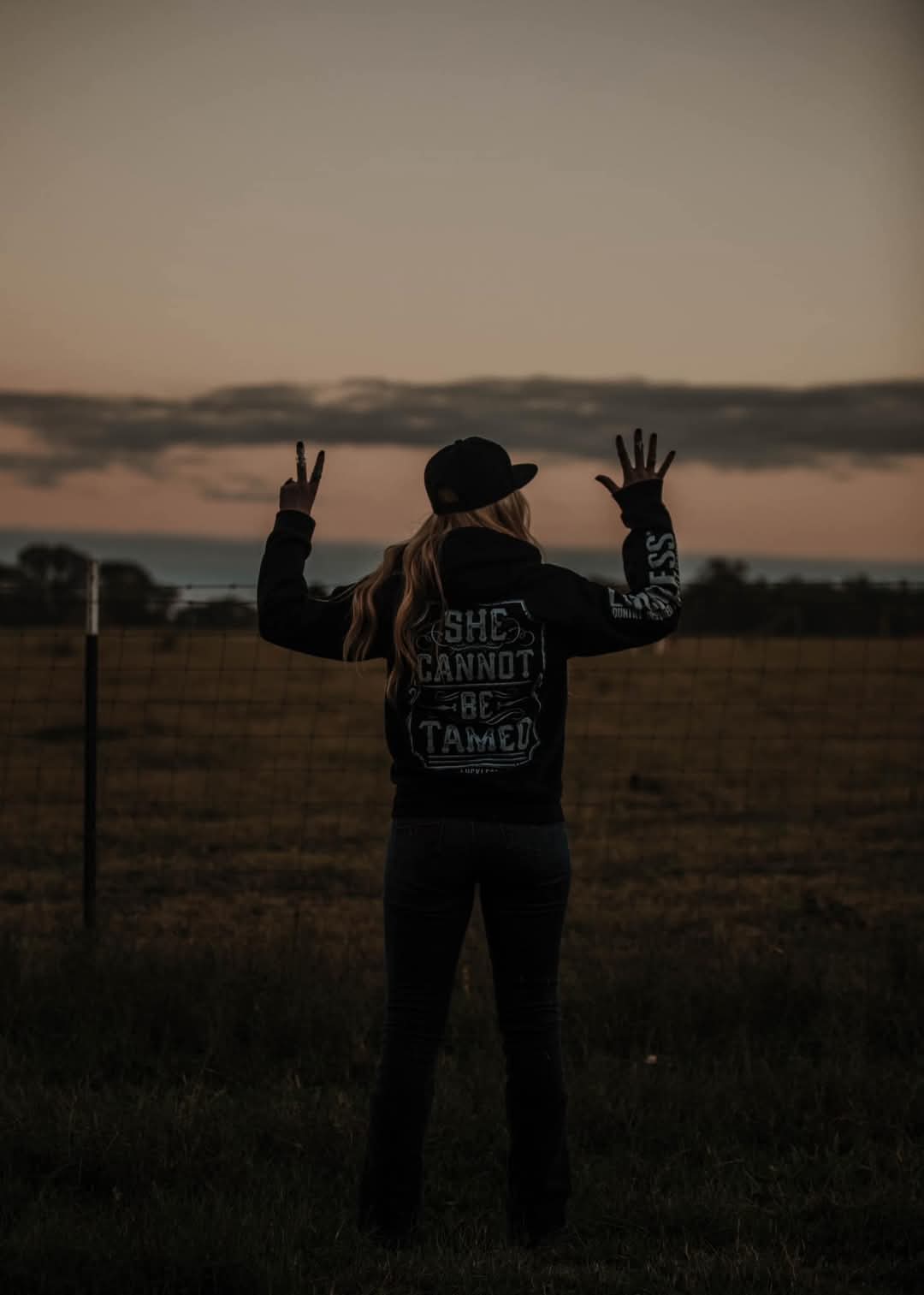 Person in a field with 'She Cannot Be Tamed' jacket at sunset