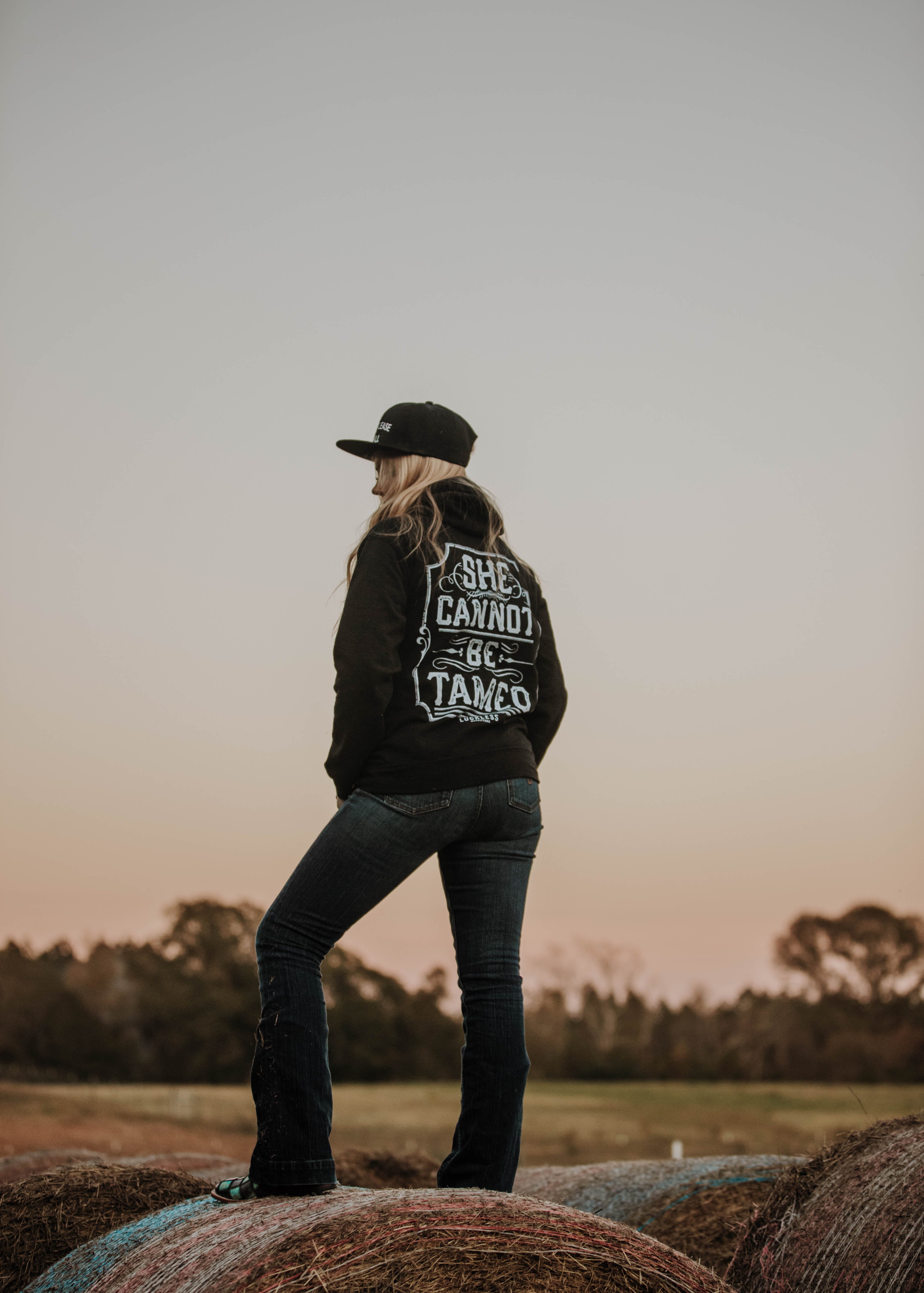 Person wearing a jacket with text standing on hay bales against a sunset sky.