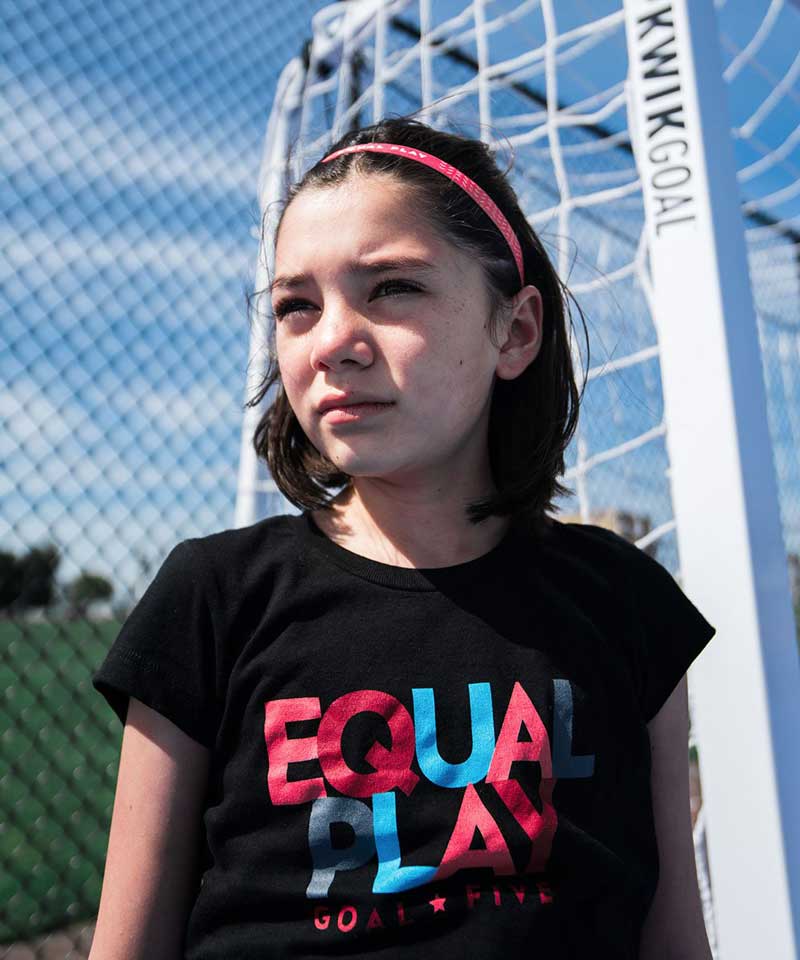 Young girl wearing a black t-shirt with 'Equal Play' text, standing in front of a goalpost.