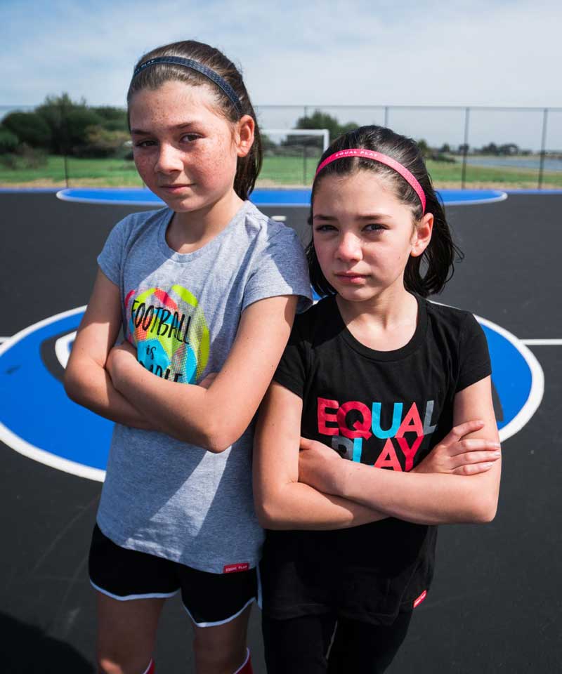 Two young girls standing on a basketball court with sports-themed shirts.