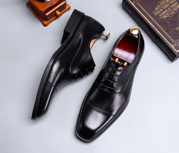 Pair of black dress shoes on a light gray background with a small book and object.