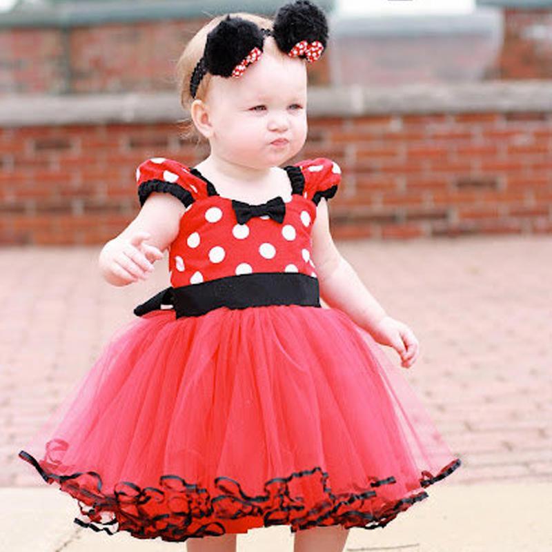Baby in a red polka dot dress with black accents standing on a brick surface.