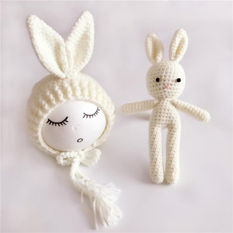 White crocheted baby bonnet with bunny ears and a matching doll on a light gray background