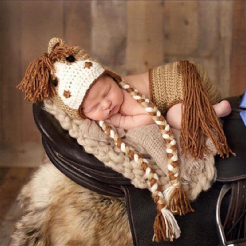 Newborn baby wearing a horse-themed knitted outfit with a hat and scarf on a wooden surface.