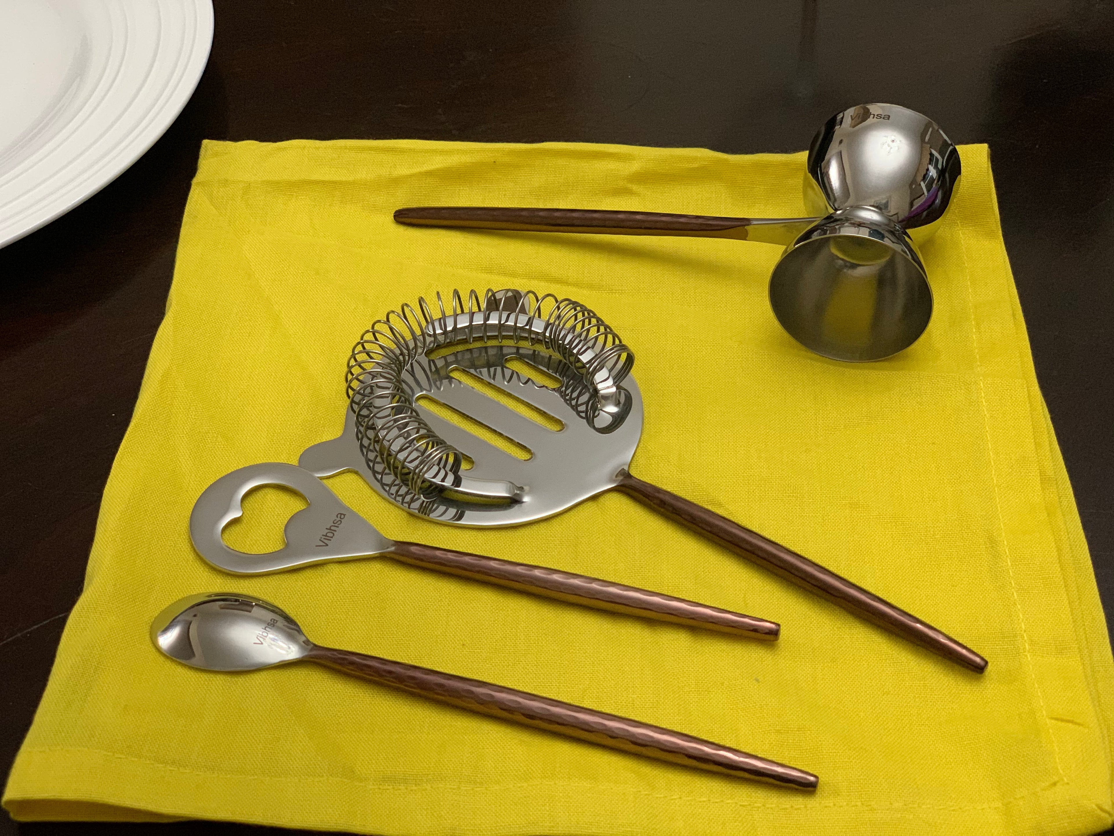 Set of bar tools including a jigger, strainer, and spoons on a yellow napkin.