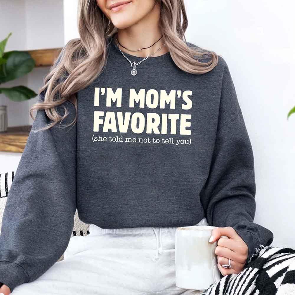 Person wearing a dark gray sweatshirt with 'I'm Mom's Favorite (she told me not to tell you)' text, holding a mug.