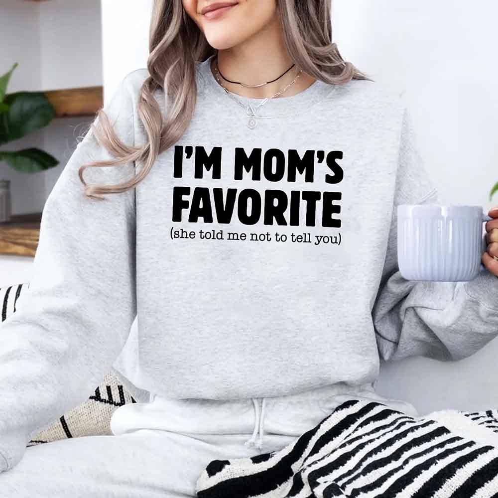 Person wearing a sweatshirt with 'I'm Mom's Favorite (she told me not to tell you)' text, holding a mug.