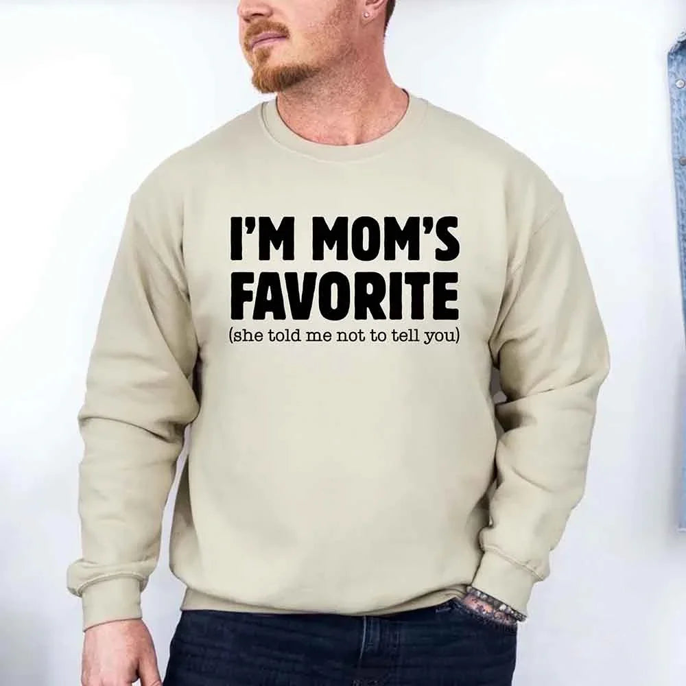 Beige sweatshirt with text 'I'M MOM'S FAVORITE (she told me not to tell you)' worn by a person.