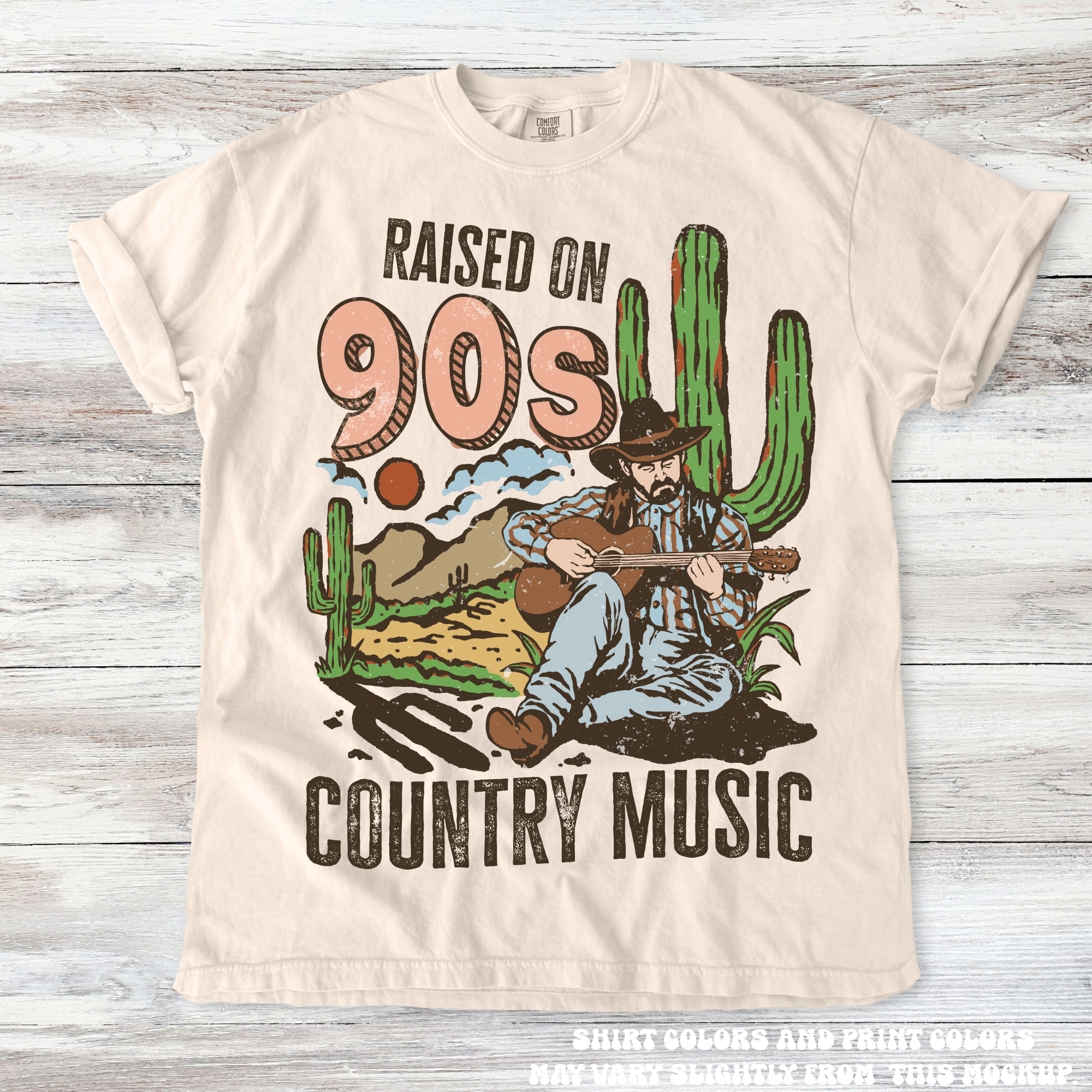 Beige t-shirt with a graphic of a cowboy playing guitar and cacti, text 'Raised on 90s Country Music' on a wooden surface.