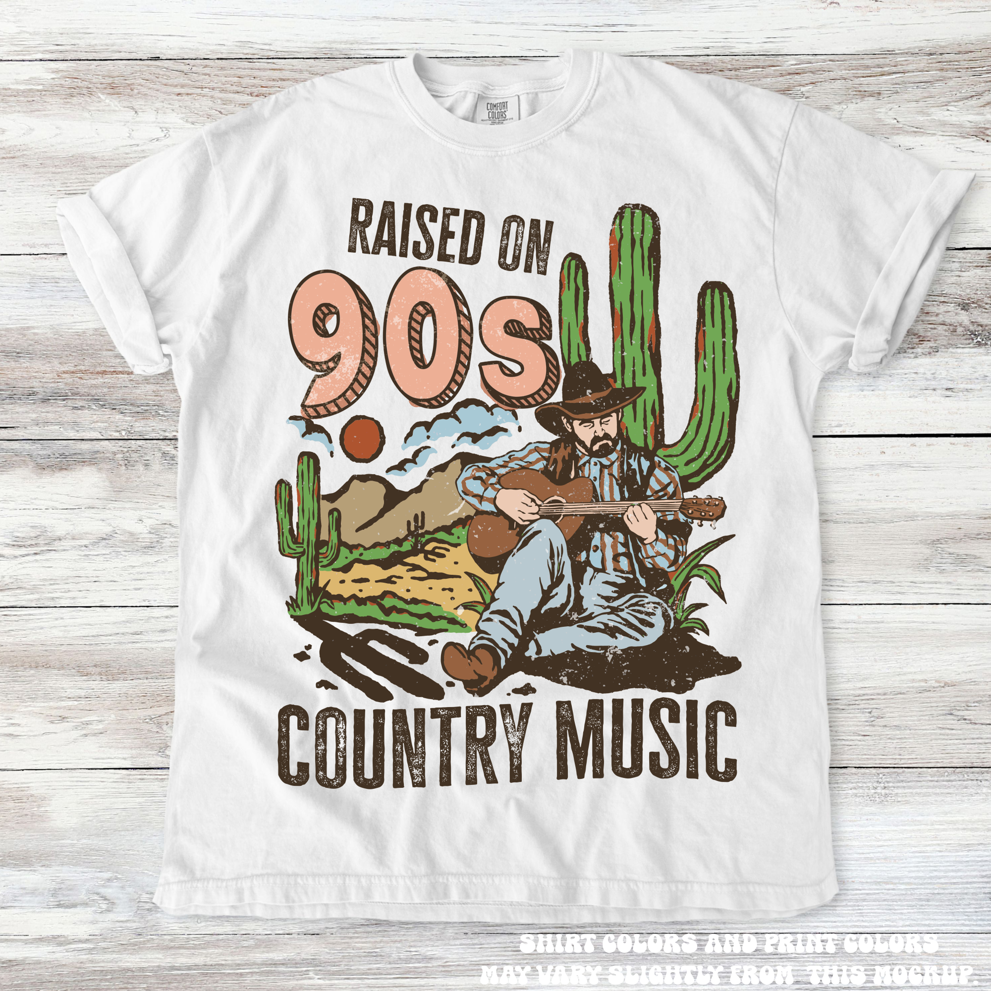 White t-shirt with a graphic of a cowboy playing guitar and cacti, text 'Raised on 90s Country Music' on a wooden surface.