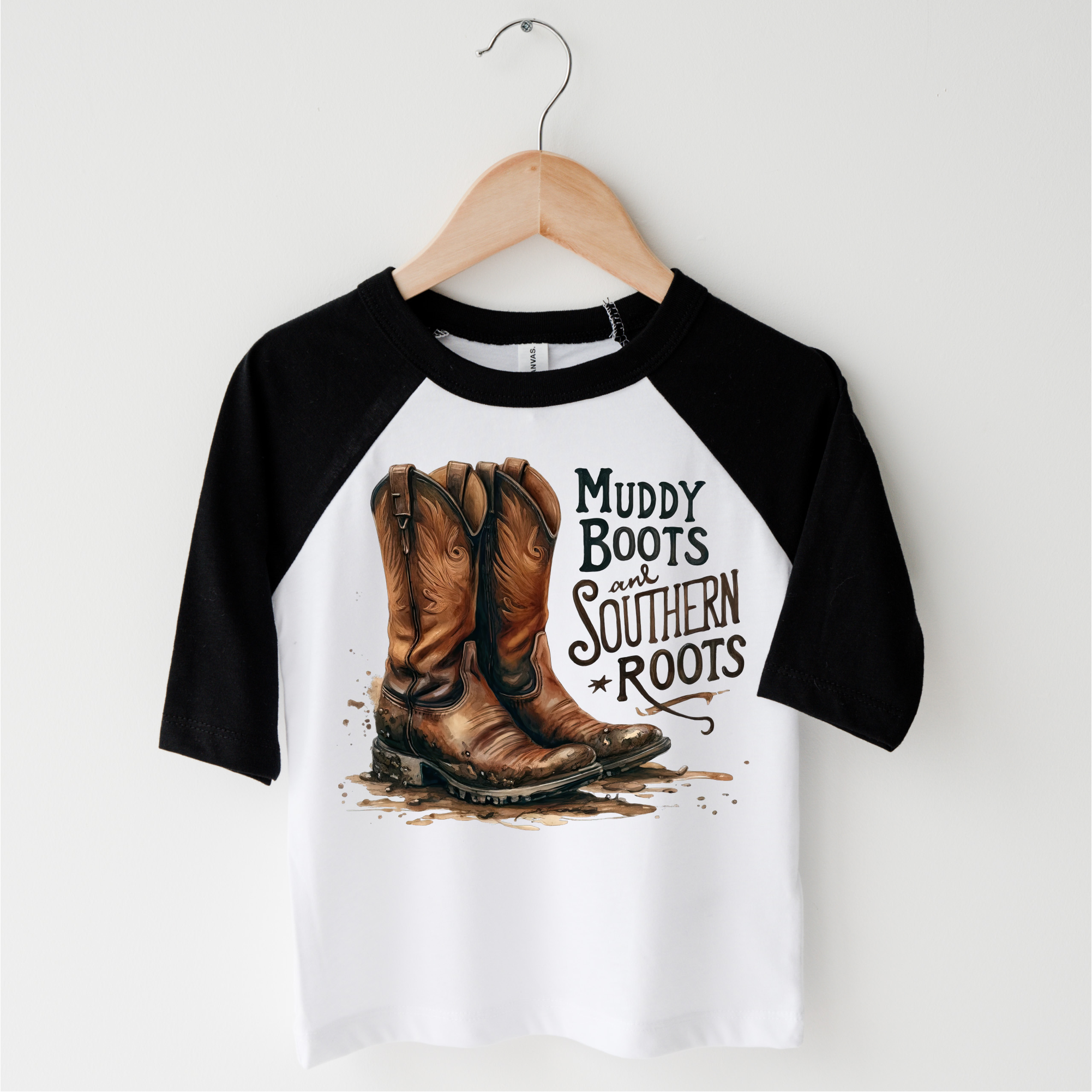 Black and white baseball shirt with cowboy boots and 'Muddy Boots and Southern Roots' text on a hanger.