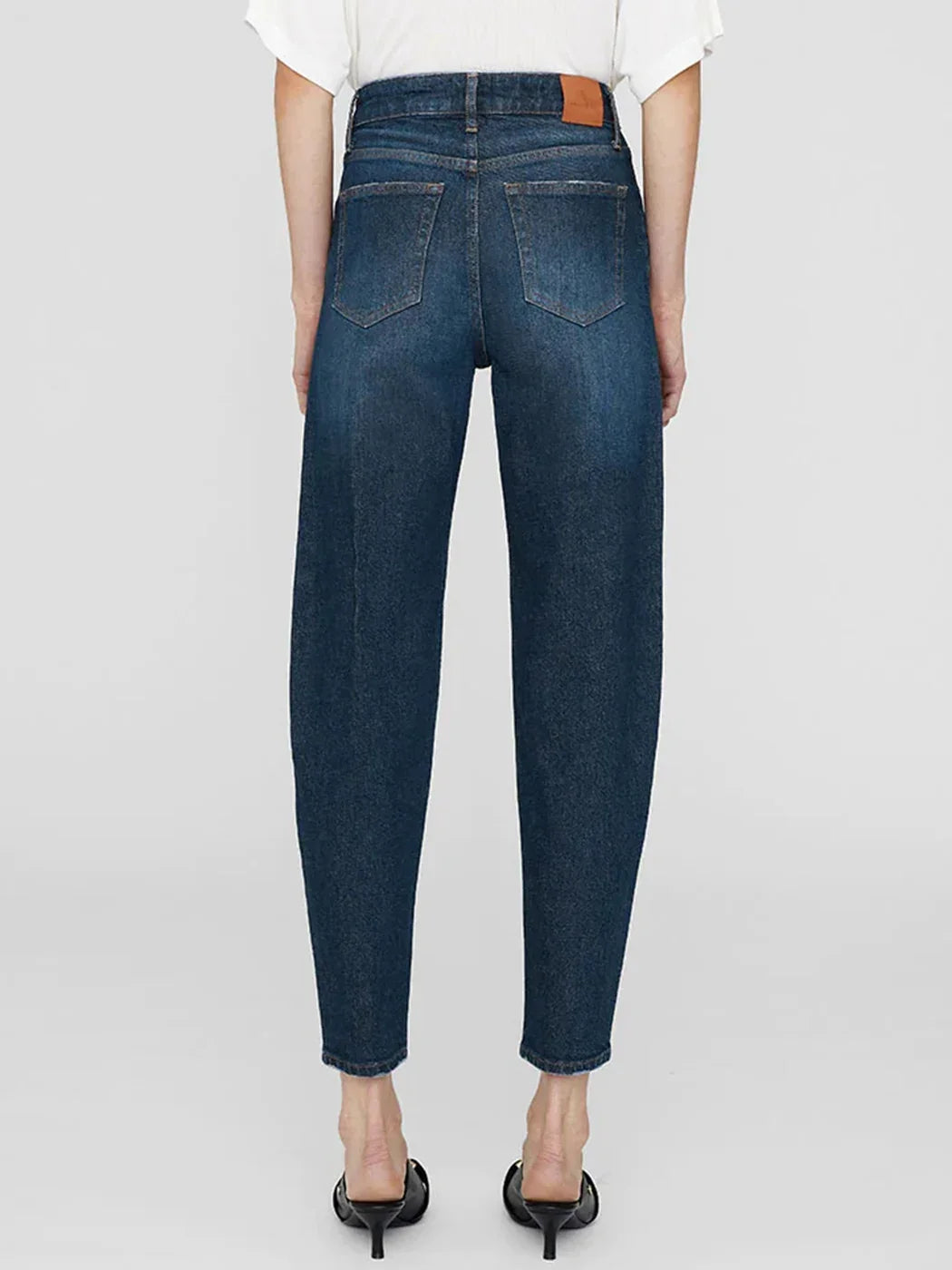 Back view of a model wearing dark blue high-waisted cropped jeans with a straight leg, paired with a white top and black heels, on a light gray background.