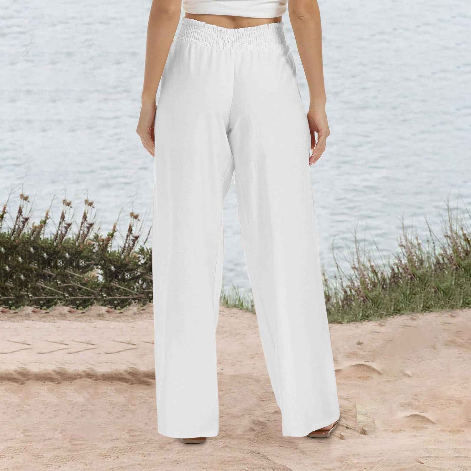 Person wearing white wide-leg pants standing on a sandy surface with a textured wall and plants in the background.