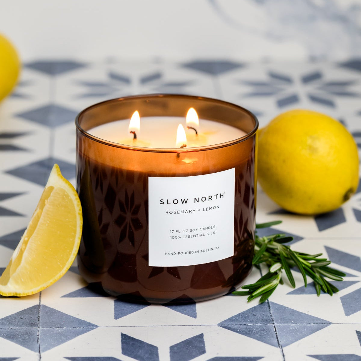 Lit candle with 'Slow North' label surrounded by lemons and rosemary on a patterned surface