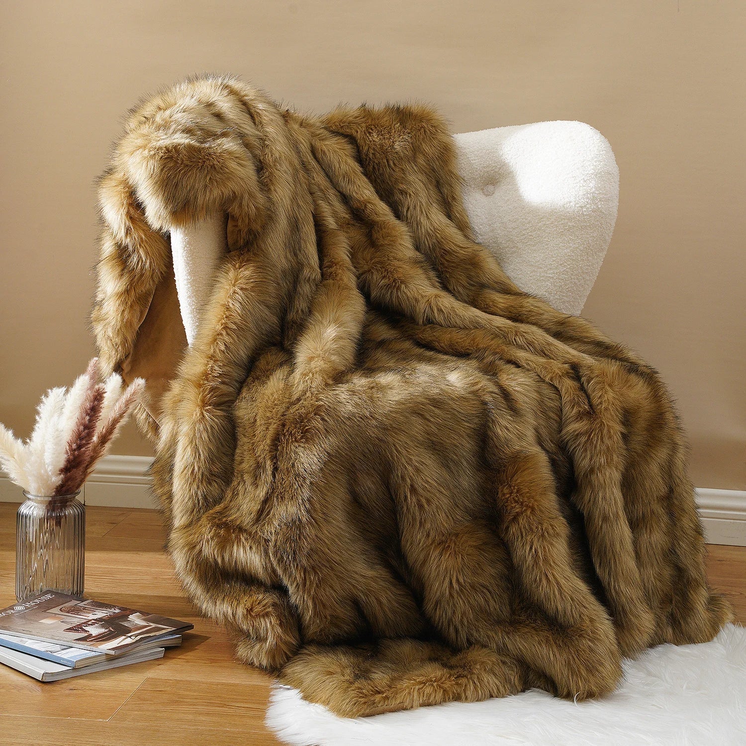 A plush faux fur throw blanket in warm brown and beige tones draped over a white armchair in a styled room setting with a wooden floor, books, and a vase.