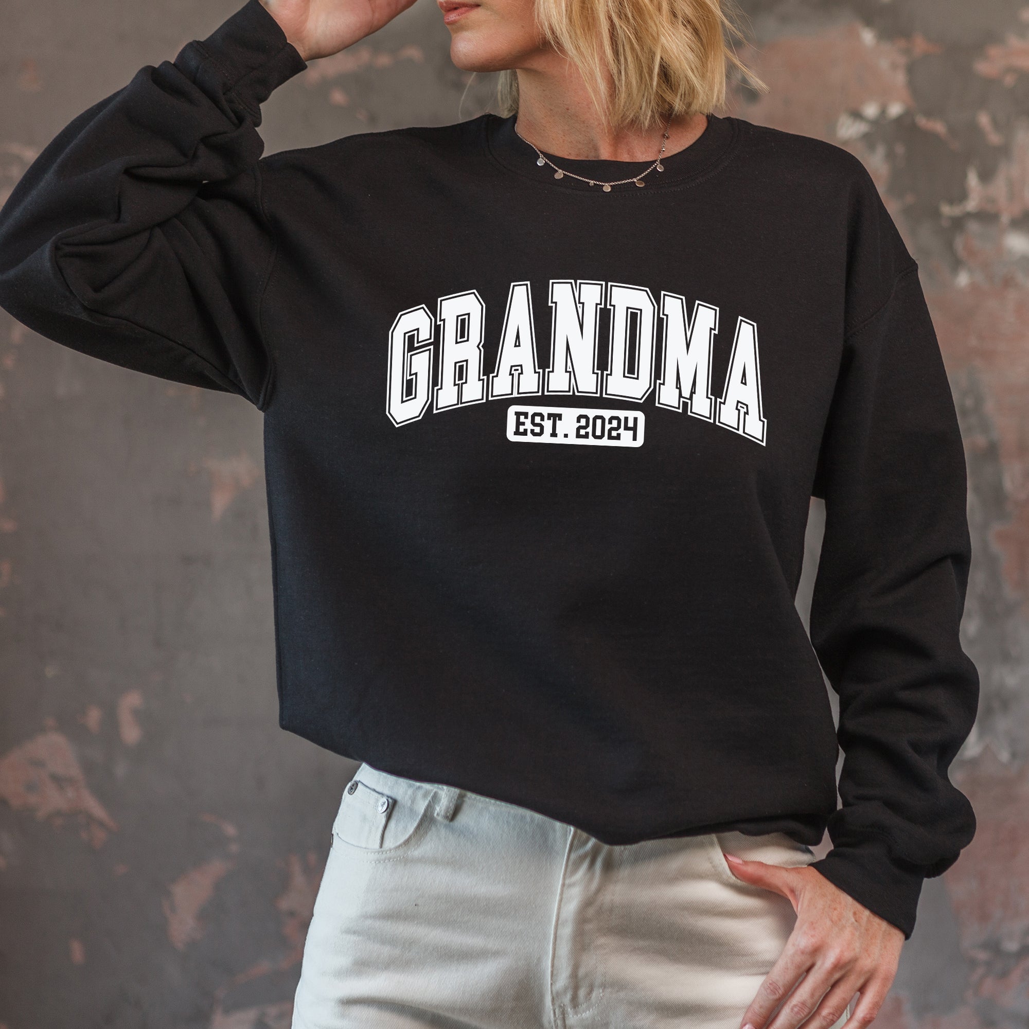 Person wearing a black sweatshirt with 'GRANDMA EST. 2024' text against a textured wall.