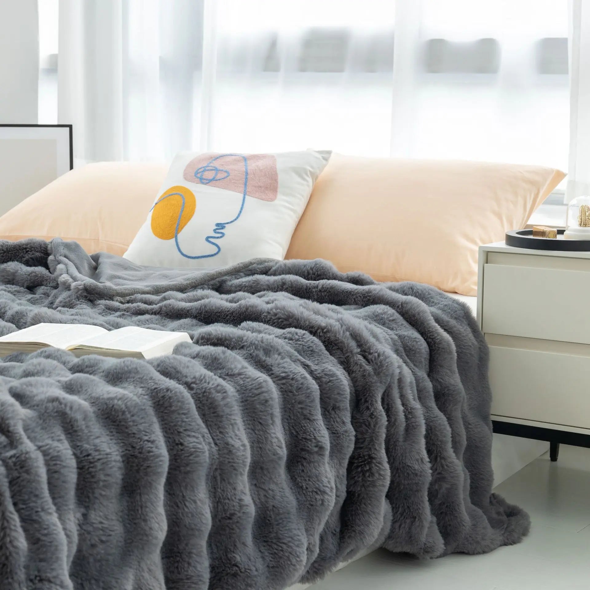 Gray fuzzy blanket draped over a bed with a pillow in a bright bedroom.