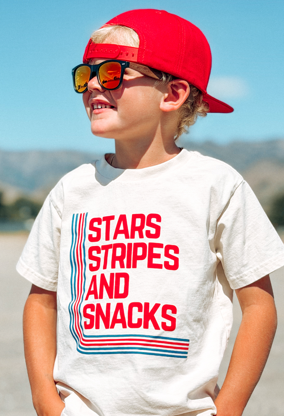 Child wearing a white t-shirt with 'Stars Stripes and Snacks' text, red cap, and sunglasses on a beach.