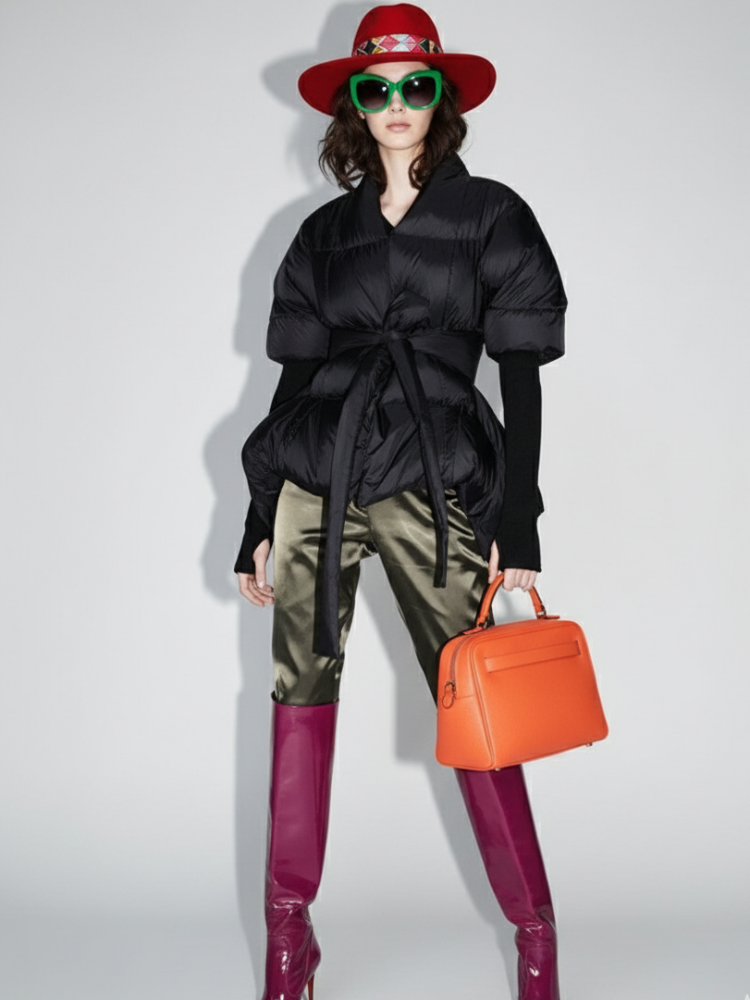 Model wearing a black puffer jacket, red hat, green sunglasses, camouflage pants, pink boots, and holding an orange bag on a white background.