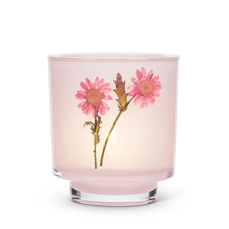 A pink glass candle jar with a clear lid, featuring two pressed pink flowers and green stems embedded on the front, set against a plain white background.