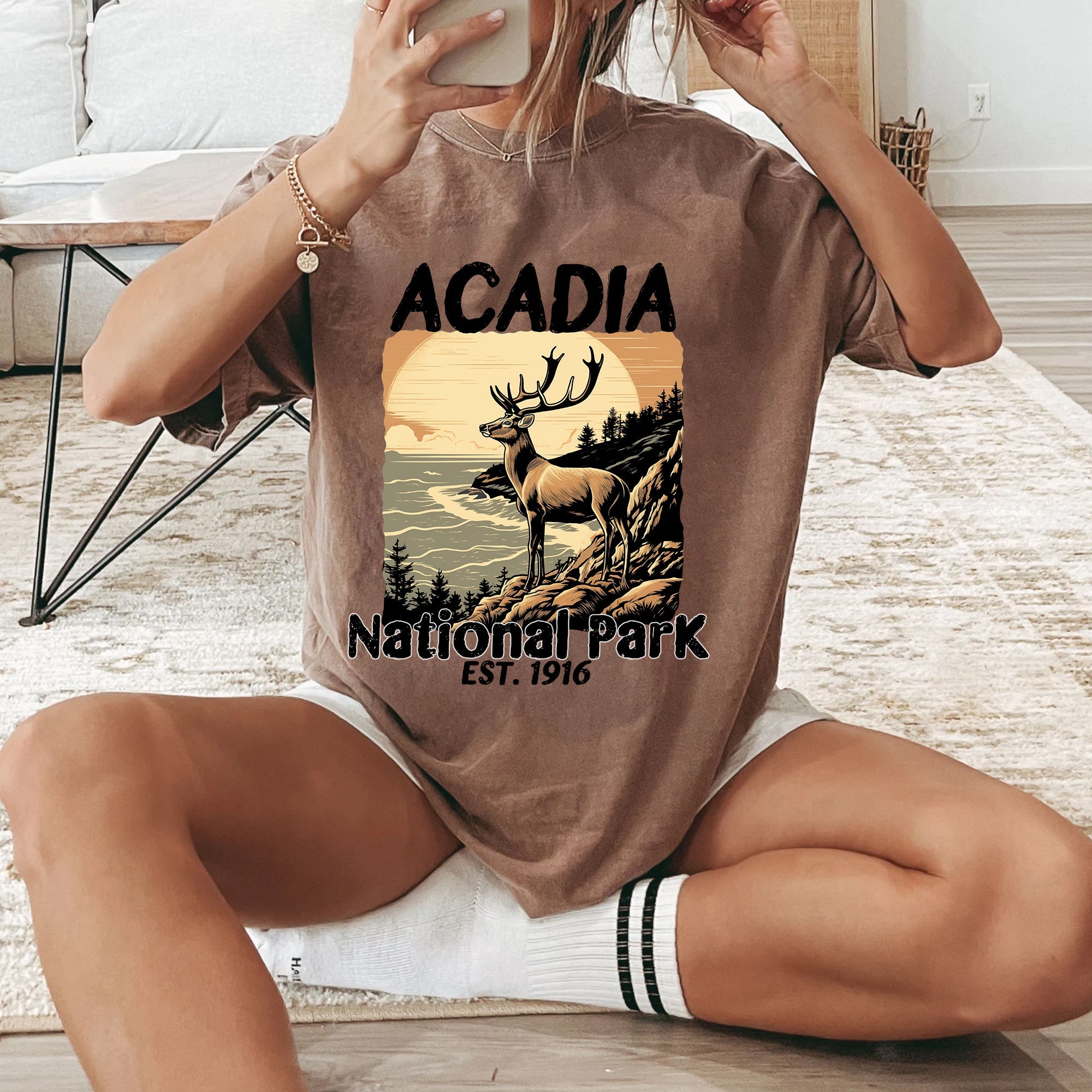 Person wearing a brown t-shirt with 'Acadia National Park' design, sitting on a wooden floor.