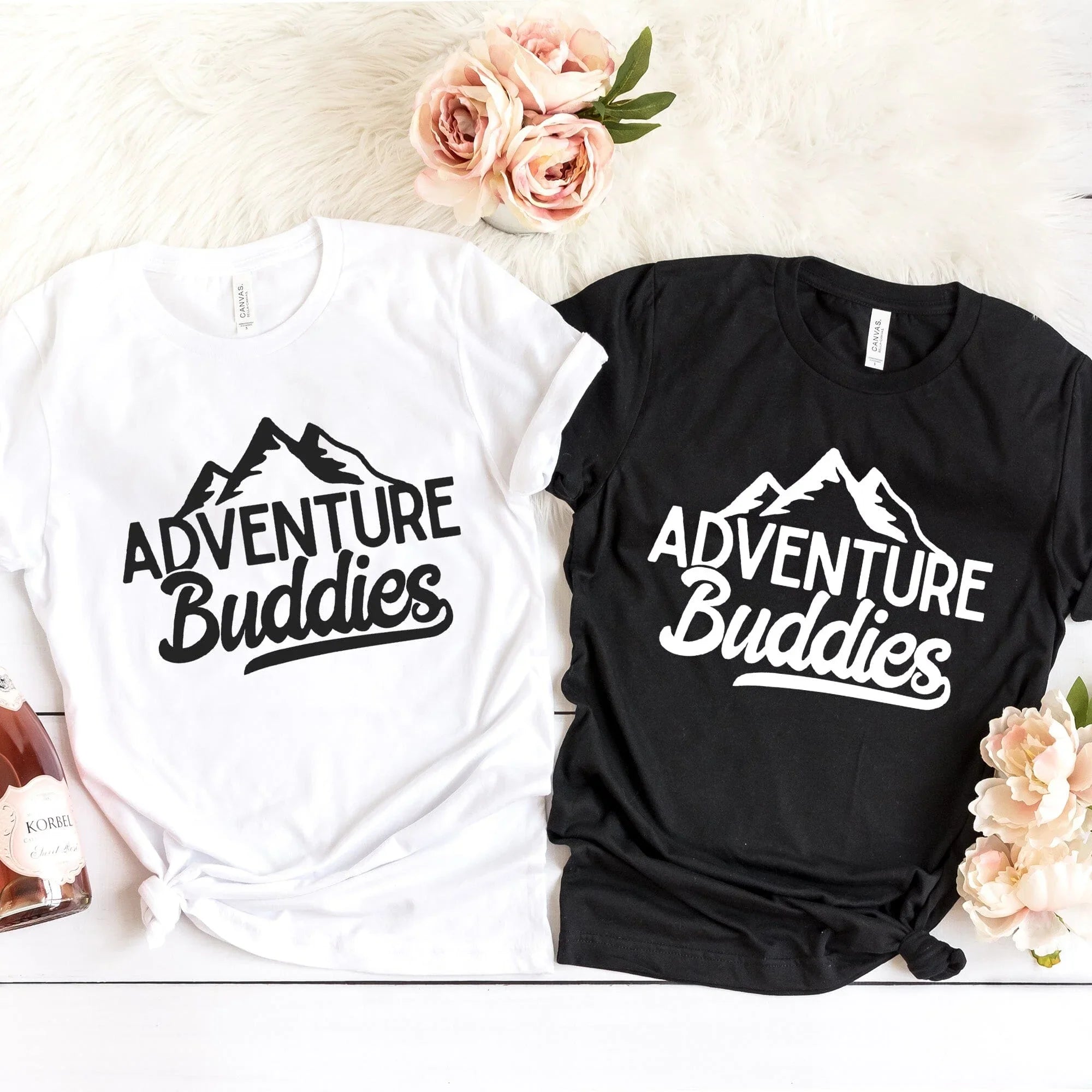 Two 'Adventure Buddies' t-shirts, one white and one black, on a light background with flowers.
