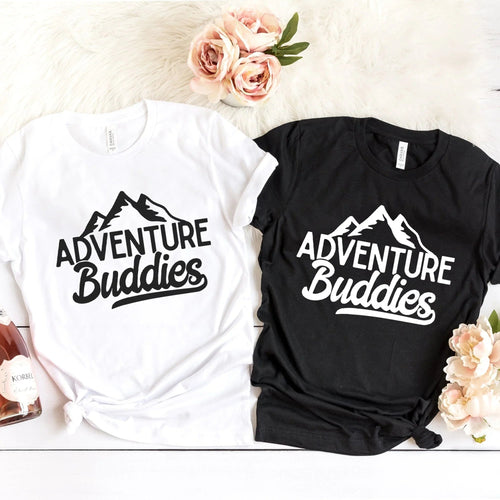 Two 'Adventure Buddies' t-shirts, one white and one black, on a light background with flowers.