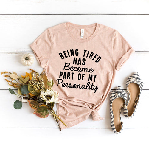 Pink t-shirt with text 'Being tired has become part of my personality' on a white wooden surface with shoes and flowers.