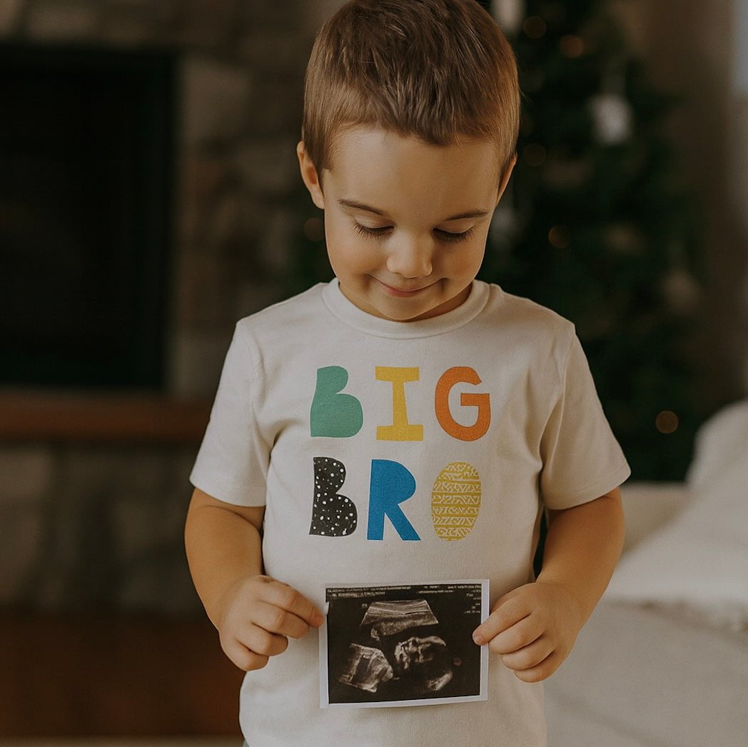 Child wearing a 'BIG BRO' shirt holding an ultrasound photo, with a Christmas tree in the background.