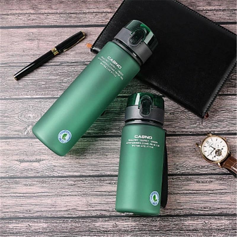 Two green water bottles with black lids on a wooden surface, surrounded by a pen, notebook, and watch.