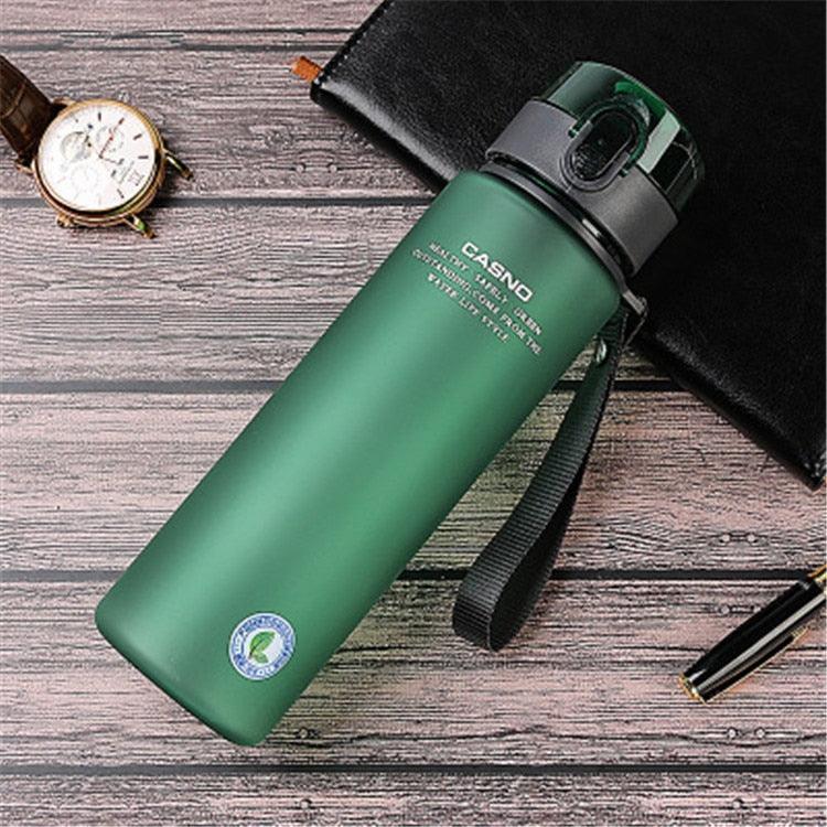 Green water bottle with black lid and strap on a wooden surface with a watch and pen.