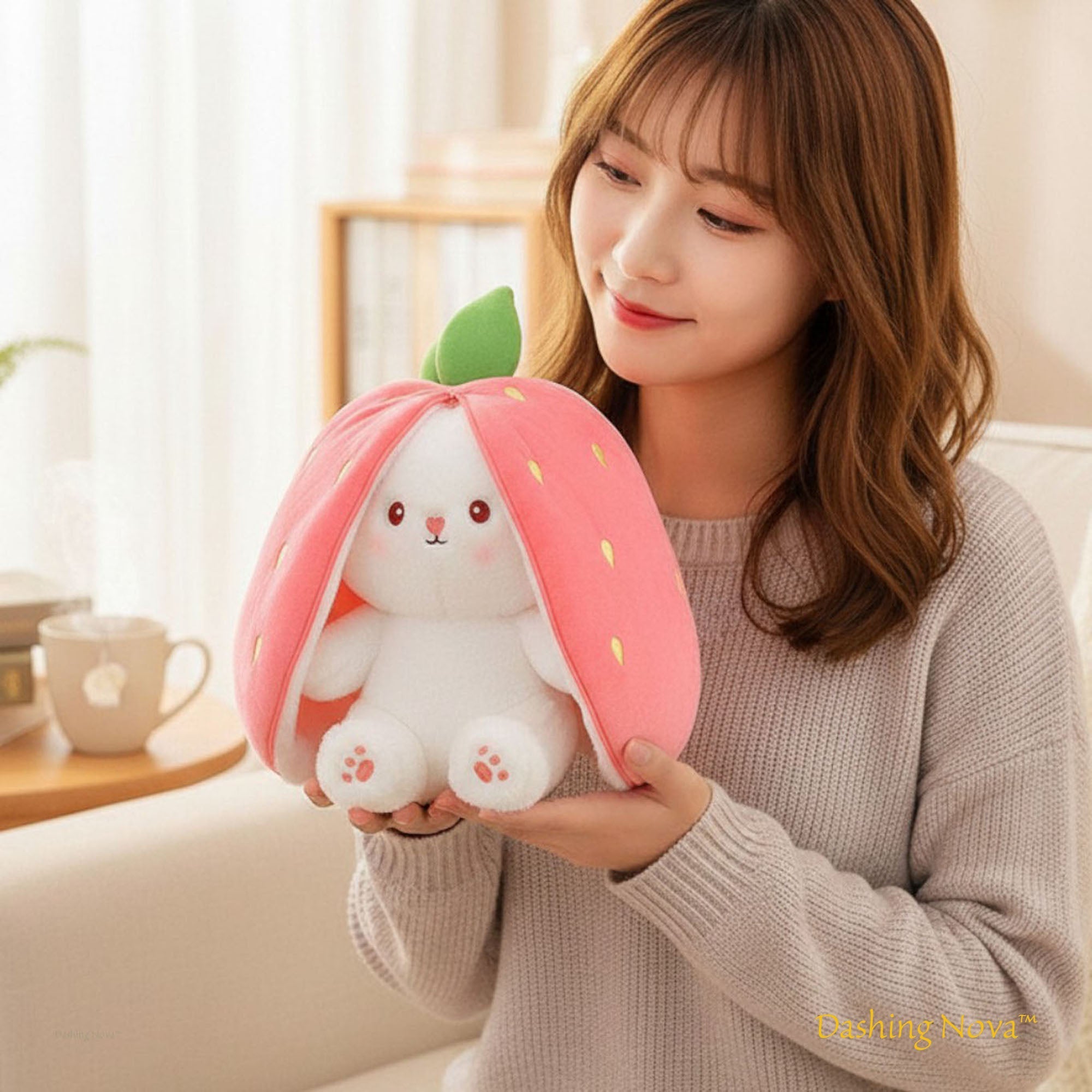 Person holding a plush toy shaped like a strawberry with a bunny design in a cozy indoor setting.