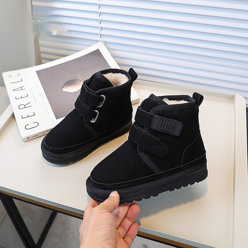 Black snow boots with a strap held by a hand on a table with a magazine in the background.