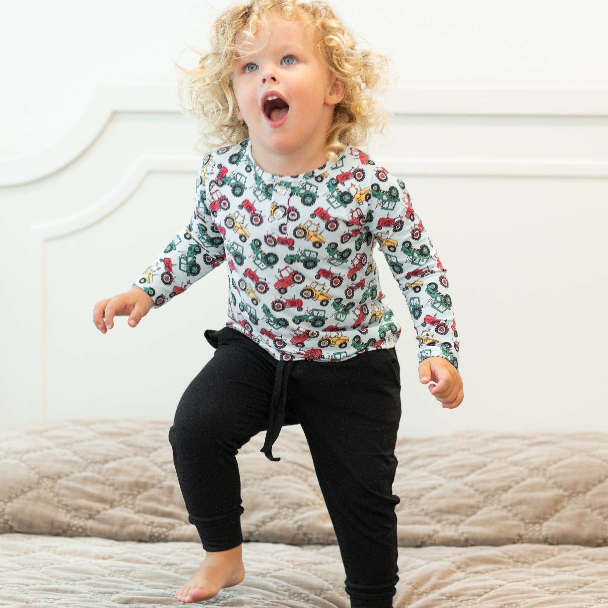 Child wearing a colorful long-sleeve shirt with vehicle patterns on a bed.