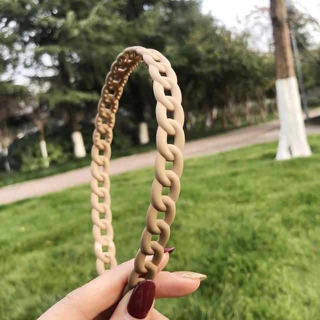 Beige braided headband held by a hand with red nail polish against a blurred green outdoor background.