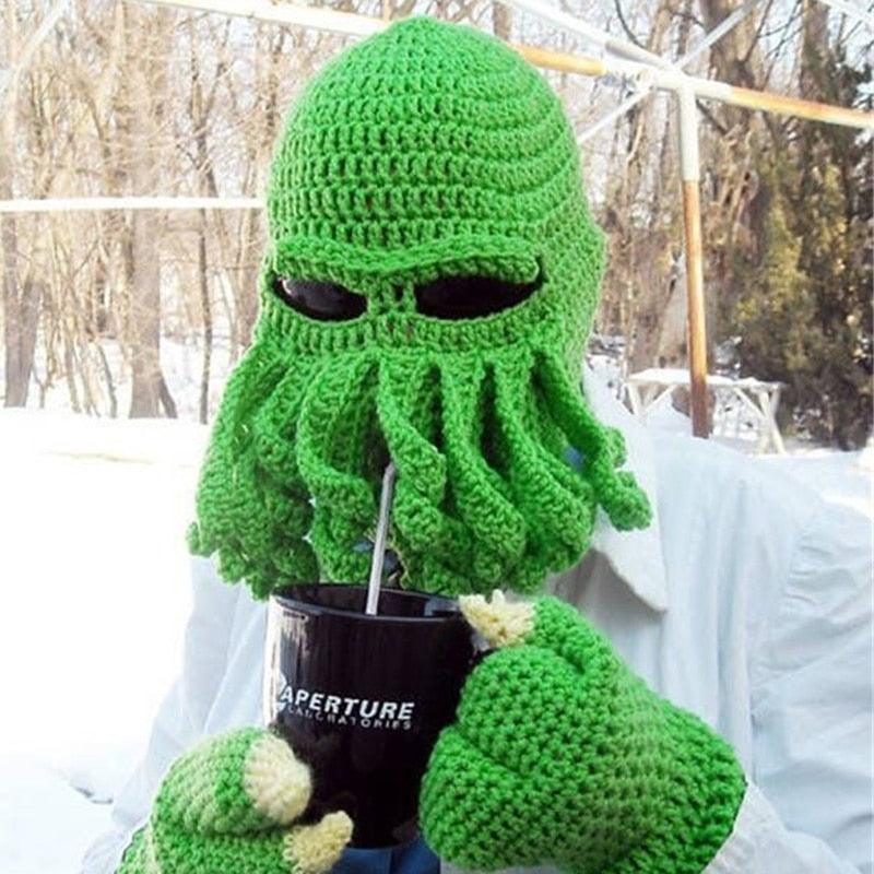 Green crochet cthulhu mask with tentacles around a black cup in a snowy outdoor setting