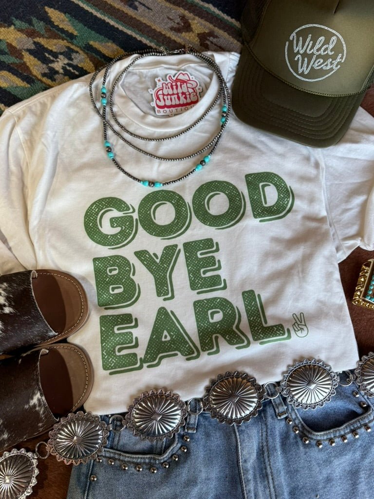 White t-shirt with 'Good Bye Earl' text, green necklace, brown shoes, and concho belt on a patterned surface.