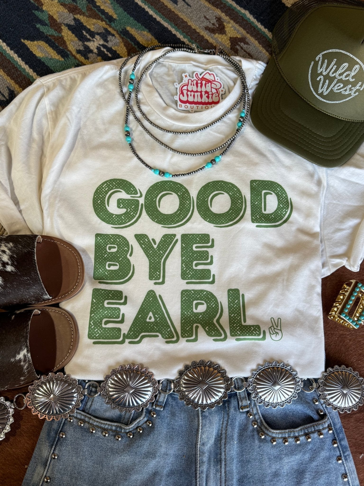White t-shirt with 'Good Bye Earl' text, surrounded by cowboy accessories on a patterned rug.